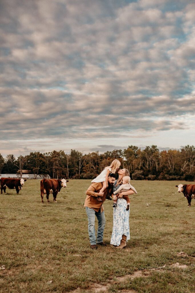 during family-photos-Pennsylvania, mom kissing daughter in cow farm on dads shoulder