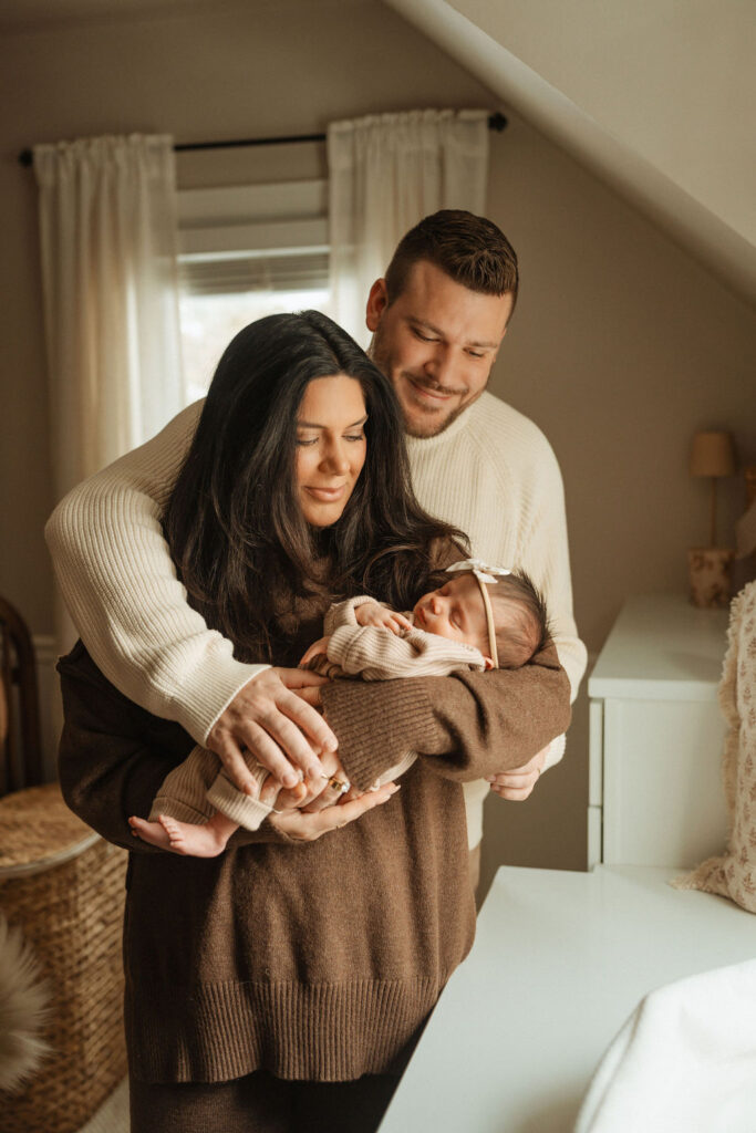 mom and dad holding newborn baby in neutral home by window